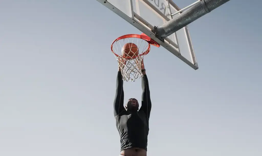Athlète effectuant un dunk puissant sur un terrain de basket en plein air sous un ciel dégagé, photographié en contre-plongée pour souligner le mouvement et la hauteur